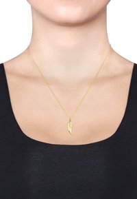 Guldkæde med en vingependant, detaljeret med indgraverede linjer. Skinnende finish, båret over en sort bluse med bådudskæring.