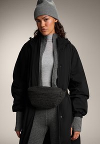 Zwarte gewatteerde jas over een grijs geribbelde trui, gecombineerd met een geweven zwarte tas en een grijze beanie, met leggings met patroon en textuur.