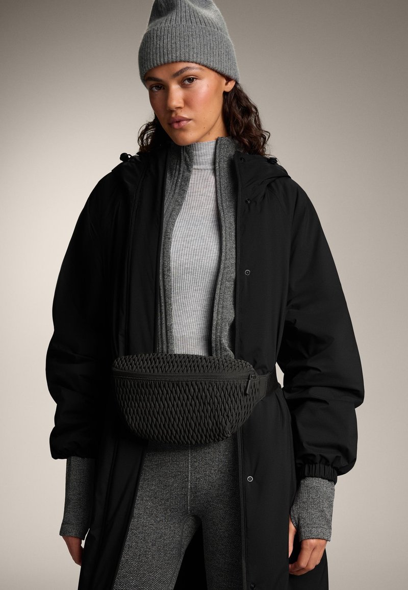 Zwarte gewatteerde jas over een grijs geribbelde trui, gecombineerd met een geweven zwarte tas en een grijze beanie, met leggings met patroon en textuur.