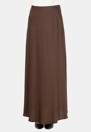 Gonna lunga, dritta e marrone indossata da una persona con scarpe nere visibili all'orlo su uno sfondo semplice.