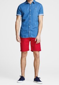 Hombre vestido con una camisa de botones azul clara y pantalones cortos rojos se mantiene de pie con confianza. Lleva anillos en los dedos y zapatillas azules.