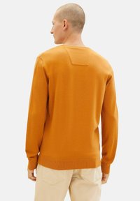 Suéter naranja de manga larga, con cuello redondo y puños acanalados. Presenta un detalle en forma de cuadrado en la parte posterior. Textura suave.