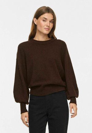 Femme portant un pull marron foncé à manches longues bouffantes et un pantalon noir, debout devant un fond blanc uni.