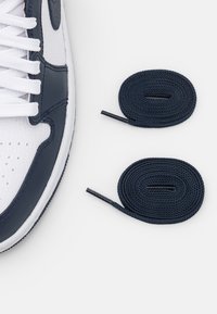 Weiße Sneaker aus strukturiertem Leder mit marineblauen Akzenten, ausgestattet mit flachen weißen Schnürsenkeln und zwei zusätzlichen flachen marineblauen Schnürsenkeln, die daneben liegen.