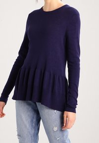 Pull bleu marine avec un col rond et des manches longues, présentant un design en peplum et des poignets côtelés. Associé à un jean délavé clair et déchiré.