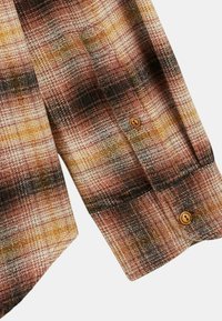 Chemise à boutons avec un motif à carreaux dans des tons de marron, jaune et noir, présentant un tissu texturé et une fermeture à un bouton au niveau du poignet.