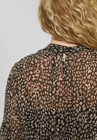 Blusa nera trasparente con stampa leopardata in tonalità beige e oro. Dettaglio a forma di chiave sul colletto e texture morbida e fluida. Drappeggio elegante.