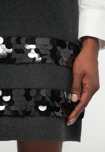 Saia cinza de lã com duas faixas horizontais de lantejoulas pretas nas bordas inferiores. As lantejoulas apresentam texturas e acabamentos variados, refletindo a luz.