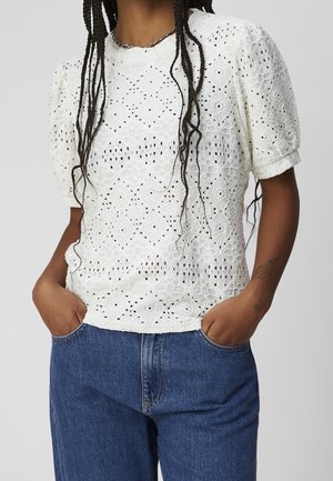 Personne portant un chemisier blanc en dentelle à manches courtes bouffantes et un jean bleu taille haute, mains dans les poches, cheveux tressés.