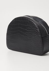 Neceser de cosméticos de imitación de cuero negro con estampado de cocodrilo, con forma redondeada y cremallera plateada. Textura suave, diseño compacto y base robusta.