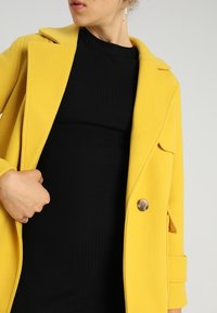 Gelber Wollmantel mit spitzem Kragen, Frontknöpfen und seitlichen Taschen, über einem schwarzen Rippenkleid kombiniert, das einen hellen Farbkontrast präsentiert.
