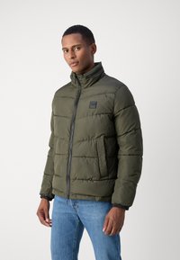 Olive green puffer jacket with a high neck, zip closure, and quilted texture; features a side pocket and black logo patch.