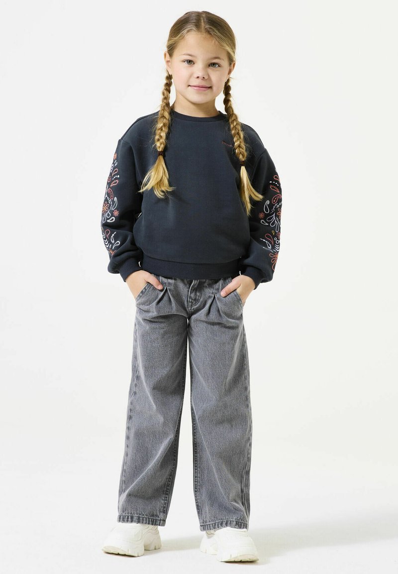 Felpa navy con ricami floreali colorati sulle maniche, abbinata a jeans grigi a gamba larga e sneakers bianche. Tessuto morbido, vestibilità comoda.