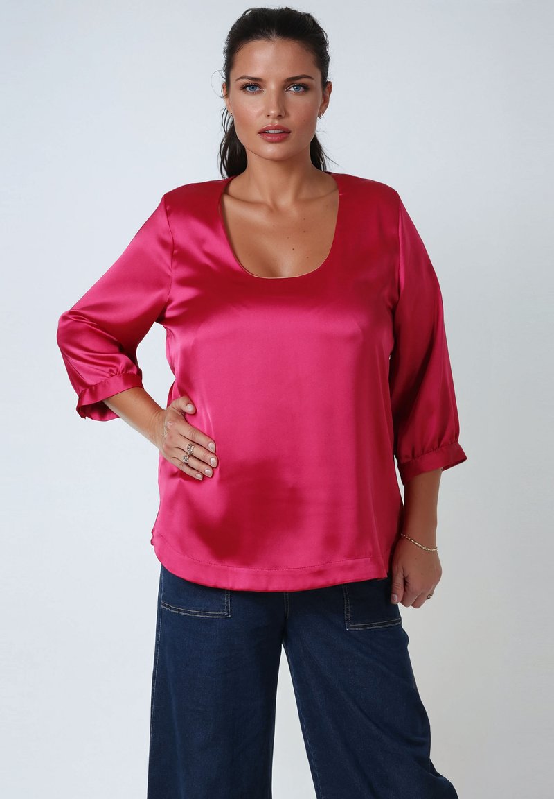 Woman with dark hair tied back wearing a shiny pink blouse with 3/4 sleeves and dark blue wide-leg pants, hand on hip against plain background.