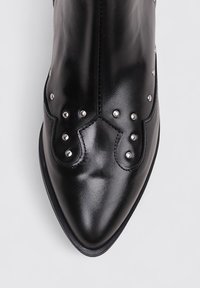 Bota negra de cuero hasta el tobillo con punta afilada, que presenta tachuelas de plata decorativas y un diseño ondulado a lo largo de la parte superior. Textura suave.