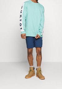 Světle tyrkysová dlouhá tričko s bílými rukávy a modrým logem, v kombinaci s námořnickými šortkami a hnědými botami se žlutými tkaničkami a šedými ponožkami.