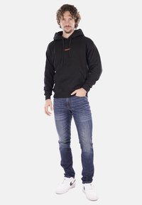 Felpa nera con cappuccio e cordini regolabili, tasca frontale e logo arancione. Abbinata a jeans blu e sneakers bianche. Look casual.
