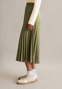 Frau, die einen langen olivgrünen, plissierten Rock, ein beiges Langarmoberteil, weiße Socken und beigetönige Turnschuhe trägt und seitlich vor einem schlichten Hintergrund steht.