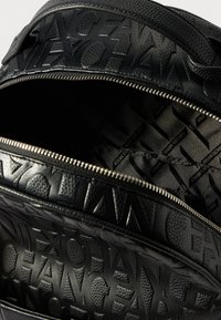 Μαύρη δερμάτινη τσάντα με υφή, με ανάγλυφο κείμενο "ARMANI EXCHANGE" και μεταλλικό φερμουάρ που είναι μισά ανοιχτό, αποκαλύπτοντας την επένδυση εσωτερικά με το λογότυπο.