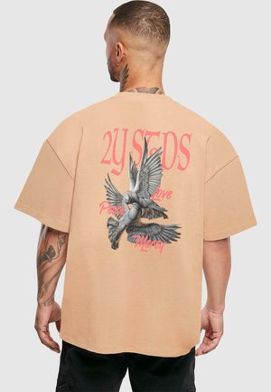Tricou bej oversized cu un grafic ce ilustrează două porumbei detaliate și textul "2Y STDS Pace Dragoste Milă" în roz. Mâneci scurte, material moale.