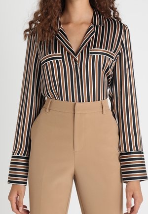 Woman wearing a brown, black, and white vertical striped blouse tucked into high-waisted beige trousers, standing against a plain background.