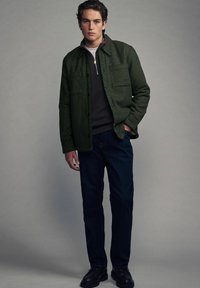Grüne strukturiertes Jacke mit zwei Brusttaschen, übereinander getragen über einem dunklen Hemd, kombiniert mit dunklen Jeans und schwarzen Schuhen. Neutraler Hintergrund.