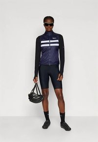 Ποδηλατική εμφάνιση με μπλε γιλέκο με λευκές ρίγες, μακριά μανίκια και μαύρο σορτς. Αξεσουάρ περιλαμβάνουν μαύρο κράνος και γυαλιά ηλίου.