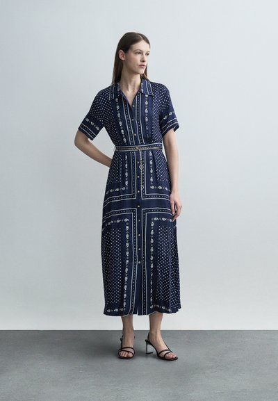 Γυναίκα που φοράει μεσοφόρεμα σε ναυτικό μπλε με λευκά σχέδια παϊέτες και κουκκίδες, κοντά μανίκια, κουμπιά μπροστά και μαύρα σανδάλια με τακούνι.