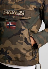 Chaqueta de camuflaje en verde, marrón y negro con un bolsillo con cremallera, que presenta un parche de la bandera noruega en rojo y una etiqueta de "NAPAPIJRI geographic".