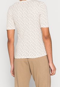 Persona con una camisa blanca ajustada de manga corta con un pequeño estampado floral marrón y pantalones beige holgados, vista desde atrás.