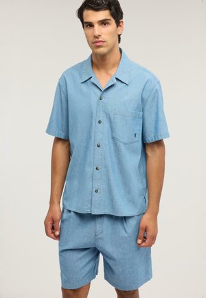 Giovane uomo con camicia azzurro chiaro a maniche corte e pantaloncini abbinati, in piedi davanti a uno sfondo semplice.