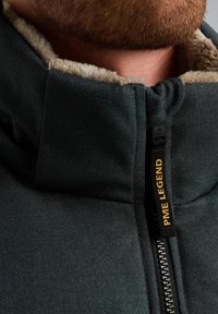 Veste noire avec un col montant, présentant une doublure en fourrure beige, une fermeture éclair à l'avant et une étiquette logo. Le tissu semble texturé et durable.