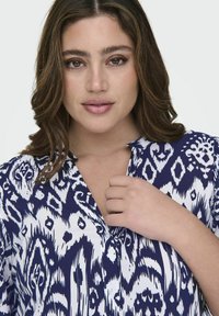 Bluza s plavim i belim vzorcem z ovratnikom in zapenjanjem na gumbe. Tkanina ima gladko teksturo in drzen abstrakten dizajn.