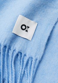 Écharpe tissée bleu clair avec des extrémités à franges et une étiquette blanche avec texte noir. Texture douce avec un motif subtil.