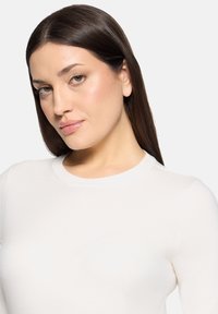 Pull à manches longues crème avec un col rond et une texture côtelée. Matière douce, coupe ajustée sur un fond neutre.