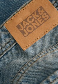 Denim material svetlo modre barve z rjavim usnjenim znakom označenim z "JACK & JONES." Ima vidne šive in mehak otip.