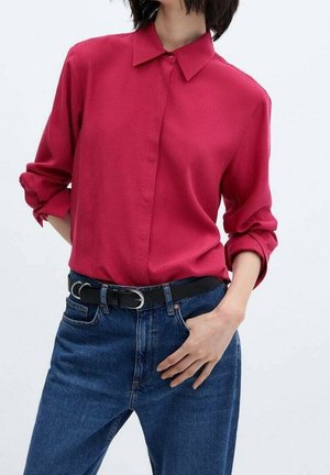 Bluzka koloru fuksji z kołnierzem i zapinana na guziki z przodu, zestawiona z wysokim stanem niebieskich dżinsów i czarnym paskiem z srebrną klamrą.