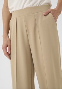 Krémové kalhoty s vysokým pasem vyrobené z hladké tkaniny, s ostrými plicemi a bočními kapsami. Diskrétní švy podél švů.