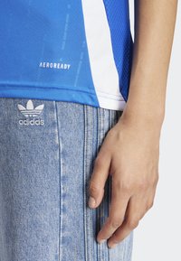 Blå sporttröja med vita detaljer och texturerad tyg, ihop med ljusblå denimbyxor med en broderad adidas-logotyp.