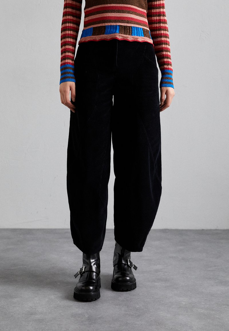 Pantalons large en velours côtelé noir avec texture visible, associés à des bottines noires à boucles. Le haut du mannequin est rayé de couleurs vives.