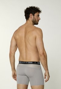 Un uomo con i capelli ricci è visto di spalle, indossa boxer grigi con una cintura nera. Lo sfondo è di un colore chiaro e neutro.