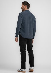 Navyblauwe corduroy shirt met een kraag, lange mouwen en een afgeronde zoom, gecombineerd met zwarte jeans en witte sneakers, gezien vanaf de achterkant.