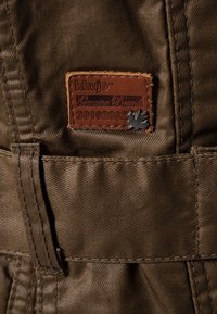Olivgrön tygdetalj med en brun läderlogotyp som har präglad text. Sömmen syns längs med sömmarna och detaljer.