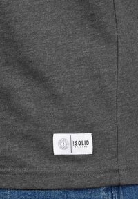 Pelēkas kokvilnas T-kreklu audums ar gludu tekstūru. I特徴 ir balts logo uz apakšējā malām, redzami šuvumi gar malām.