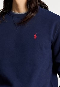 Mörkblå sweatshirt i mjukt tyg, med rund halsringning, synlig sömnad och en liten röd polo-logga på vänster bröst.