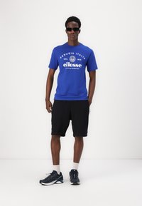 T-shirt de algodão azul com logótipo e texto, calções pretos, sapatilhas escuras e meias brancas. Modelo a usar óculos de sol, de pé contra um fundo simples.