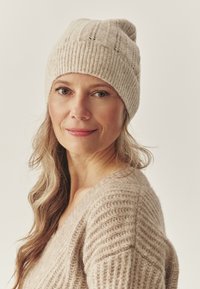 Beige knitted hat with a ribbed texture, featuring a rolled brim. Worn with a matching beige knitted sweater. Soft and cozy appearance.