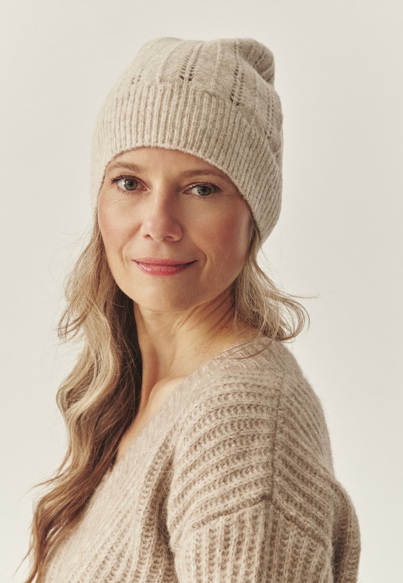 Beige knitted hat with a ribbed texture, featuring a rolled brim. Worn with a matching beige knitted sweater. Soft and cozy appearance.