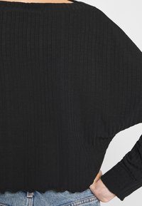 Black, ribbed sweater with a wide neckline and scalloped hem. Long sleeves and a relaxed fit. Texture is soft with subtle lines.