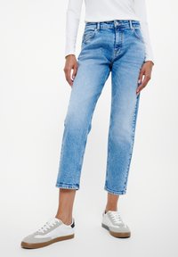 Person som bär ljusblå croppade jeans, vit långärmad topp och vita sneakers med grå och svarta accenter, stående på vit bakgrund.